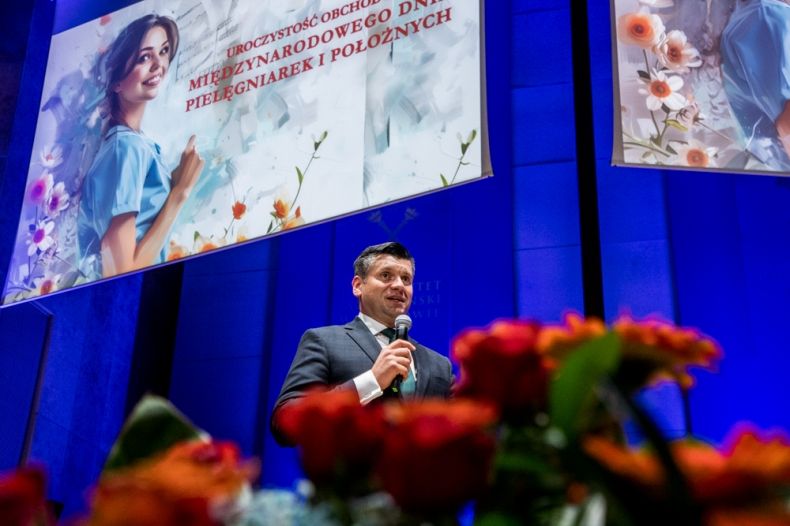Wicemarszałek Łukasz Smółka stoi na scenie, w ręce trzyma mikrofon. Na pierwszym planie widoczne czerwone kwiaty, za nim barwna plansza z wizerunkiem pielęgniarki.